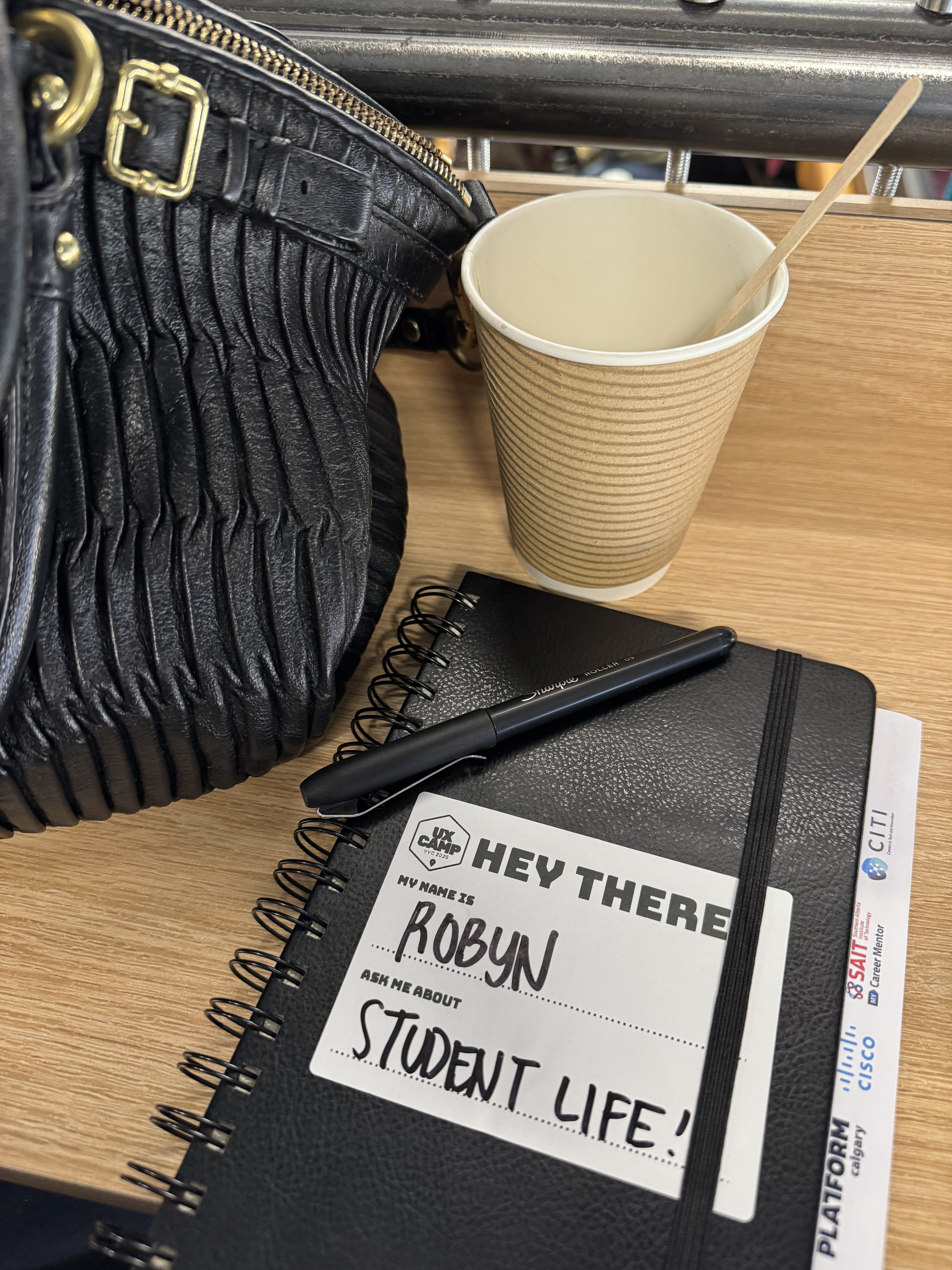 Robyn's notebook on a table at UX Camp, with a sticker on the front that reads My name is Robyn, ask me about student life